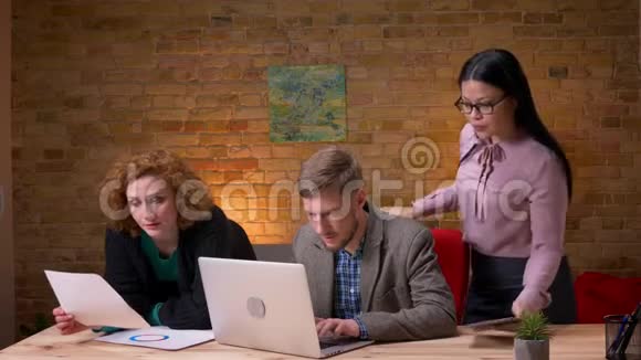 在笔记本电脑上工作并与两名女性员工讨论数据的年轻白种人商人的特写镜头一个视频的预览图