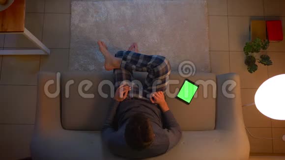 年轻男性看电视的特写镜头并使用带有绿色屏幕的平板电脑上的应用程序作为遥控器来转动视频的预览图