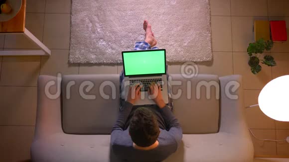 在舒适的家中坐在沙发上的年轻人用绿色屏幕在笔记本电脑上发信息的特写镜头视频的预览图