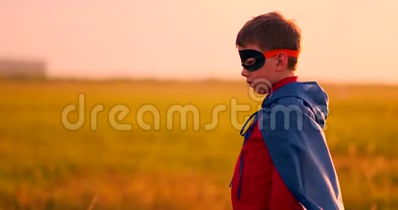一个男孩戴着超级英雄的面具看着日落时站在田野里的摄像机的肖像视频的预览图