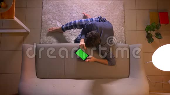 一个年轻人坐在室内的地板上看着绿色屏幕的平板电脑广告的特写镜头视频的预览图