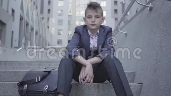 穿着考究的可爱男孩坐在街上的楼梯上这个男孩很累想做个孩子儿童视频的预览图