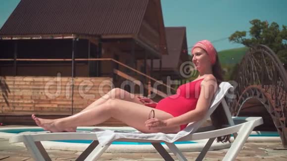 一个穿着红色一件泳衣的漂亮怀孕女孩躺在游泳池旁的休息室里那个女孩戴太阳镜视频的预览图