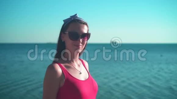 一个穿着红色一件泳衣和眼镜的怀孕女孩站在海边对着镜头微笑视频的预览图