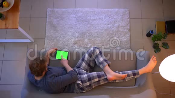 年轻的穿着随意的男性躺在沙发上用绿色屏幕在平板电脑上播放视频的特写镜头视频的预览图