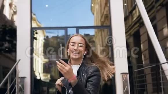 戴眼镜的漂亮女孩在戴耳机时使用智能手机应用程序听音乐视频的预览图