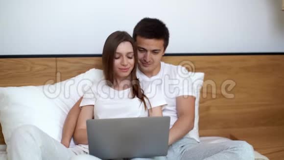 一对漂亮的年轻夫妇在家里躺在床上时用笔记本电脑和微笑视频的预览图