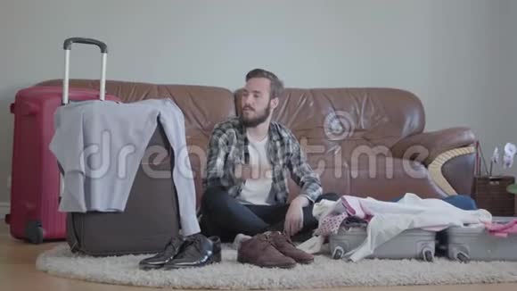 英俊的糊涂的长胡子的男人坐在地板上家里靠近皮沙发前的手提箱很多衣服视频的预览图