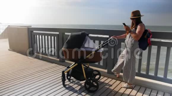 一位年轻的母亲站在木板路上带着一辆婴儿车用手机拿着自拍全垒打视频的预览图