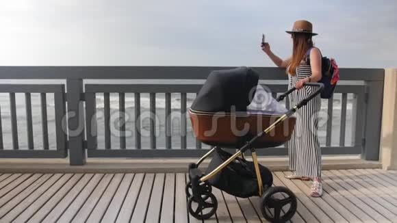 一位年轻的母亲带着一辆电车在木板路上散步并在电话中拍下了大海的照片全垒打视频的预览图
