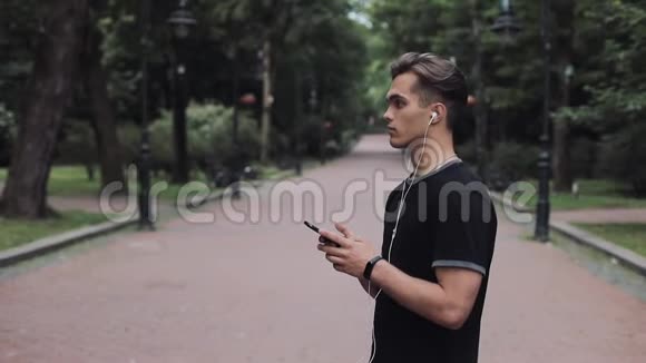 波特拉尔英俊的年轻人戴着耳机和智能手机手里拿着健身追踪器站在公园里视频的预览图