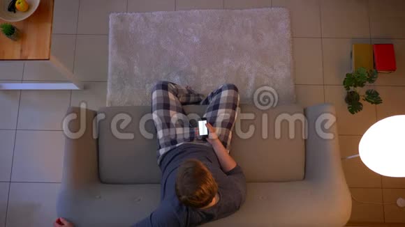 年轻的穿着随意的男性坐在沙发上看电视用手机上的应用程序作为特写镜头视频的预览图