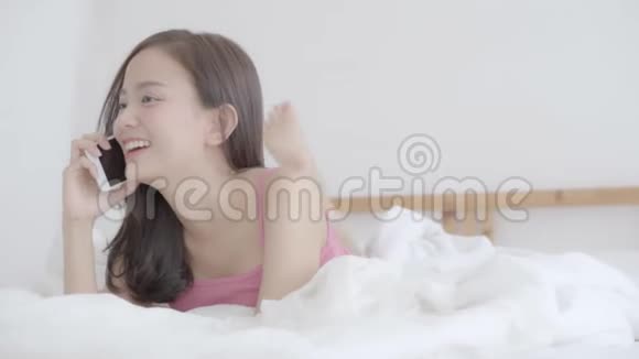 美丽的肖像年轻的亚洲女人微笑着躺着说着智能手机在卧室里放松视频的预览图