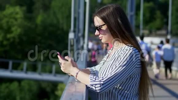 戴太阳镜的年轻女孩在旅游桥上使用智能手机视频的预览图