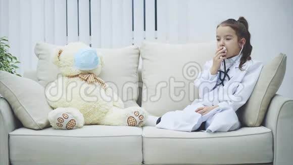 穿着医用外套的小女孩在听诊器里说话戴着医用面具的白色泰迪熊正坐在她身边视频的预览图