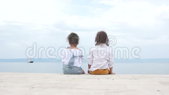 男孩和一个女孩坐在海边看着远处的一艘帆船旅费视频的预览图