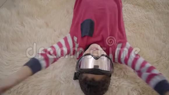 特写一幅可爱的非裔美国男孩躺在米色蓬松地毯上的肖像他的滑雪护目镜视频的预览图