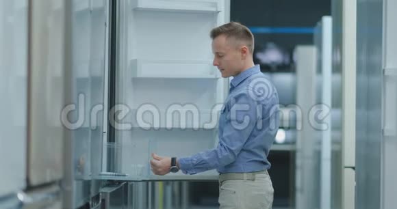 一个穿着蓝色衬衫的年轻人打开电器商店冰箱的门并与其他型号进行比较视频的预览图