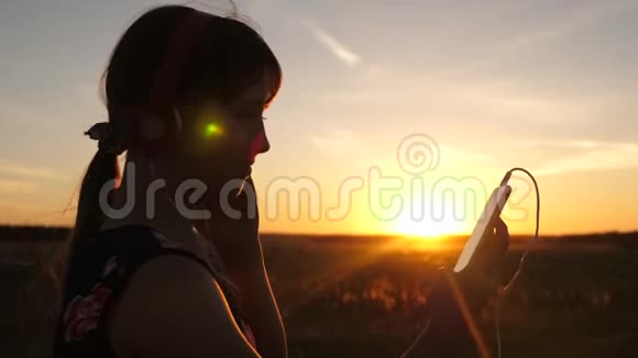快乐的女孩在美丽的夕阳下在你的智能手机上旅行和听音乐公园里的小女孩视频的预览图