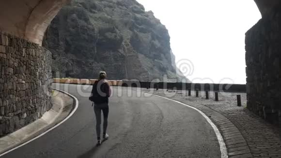 镜头跟随一名女子走在一条路上背着背包从一条隧道走向海洋上方的悬崖边缘举起手臂视频的预览图