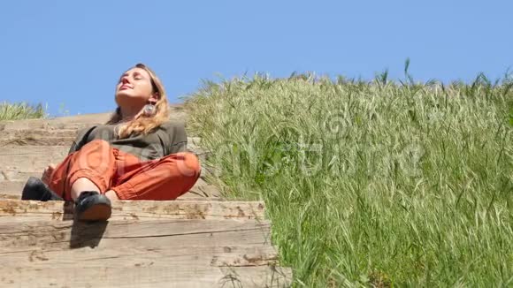 年轻的白种人女人坐在木楼梯上享受阳光下的生活视频的预览图
