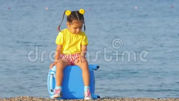 穿着五颜六色衣服的小可爱女孩坐在海边的蓝色手提箱上视频的预览图