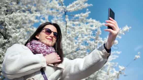美丽的微笑女人自拍使用智能手机摆姿势白色开花樱花树背景视频的预览图