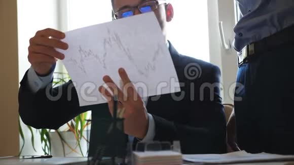 年轻英俊的戴眼镜的商人手里拿着一张带有统计图的纸为男性提供指导视频的预览图