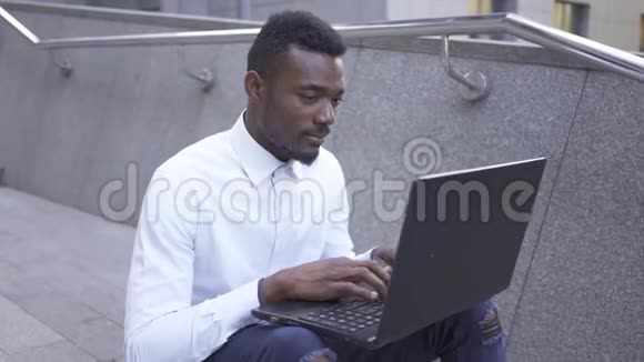 积极的非裔美国人穿着白色衬衫坐在城市街道上的笔记本电脑上打字自信的男性商人或视频的预览图