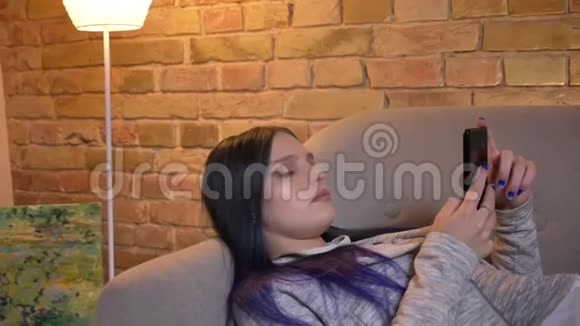 一个舒适的公寓里躺在沙发上用电话上网的年轻白种人时髦女性特写镜头视频的预览图