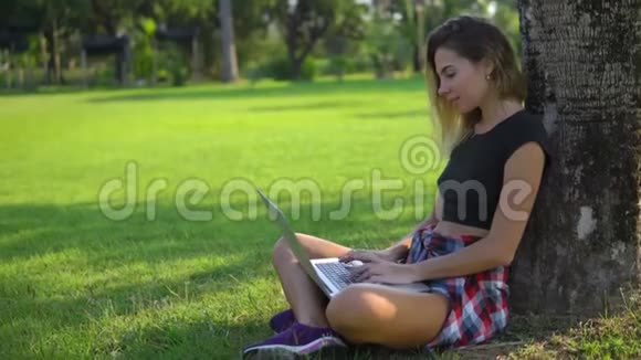 漂亮的成年女孩坐在公园的树下把电脑放在膝盖上视频的预览图