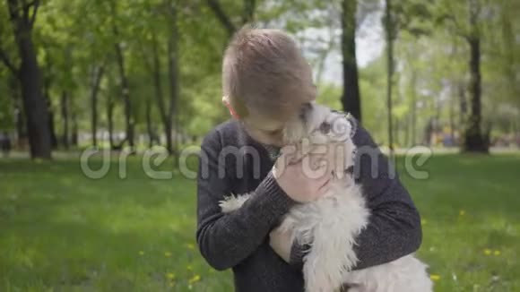 肖像可爱的小男孩抱着一只白色的毛茸茸的狗在美丽的绿色公园里视频的预览图