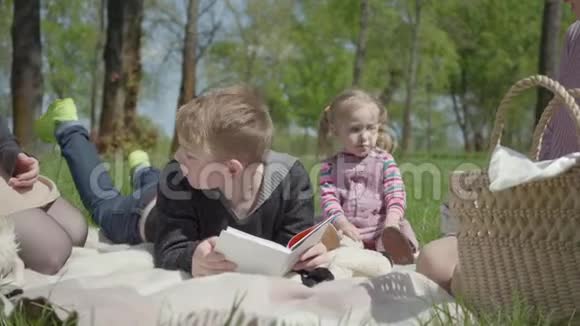 十几岁的金发男孩躺在公园的毯子上看书可爱的小女孩穿着粉红色的裙子坐在附近二视频的预览图