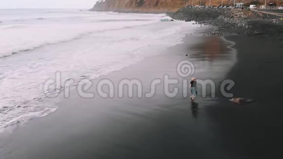 女子漫步沙滩鸟瞰穿着时尚的女孩在黑色火山海滩电影顶部的景色视频的预览图
