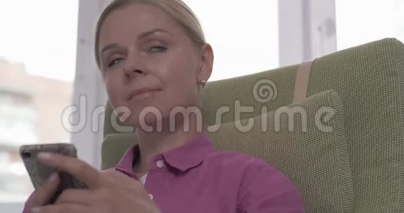 漂亮的金发女孩坐在扶手椅上在智能手机上发短信视频的预览图