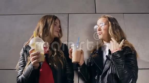 两个美丽的快乐女孩一边说话一边喝着鸡尾酒一边站在现代建筑旁边的灰色墙壁附近户外活动视频的预览图