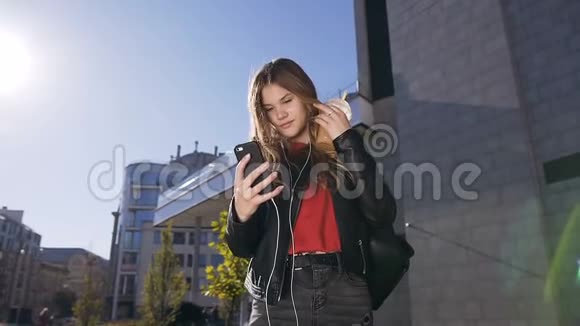 年轻貌美的女士戴着长发耳机在城市街头散步时使用智能手机视频的预览图