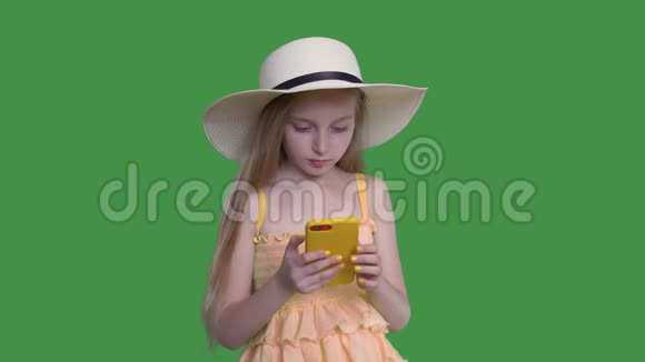 戴着夏帽的小女孩正在绿色墙背景上浏览智能手机阿尔法频道视频的预览图