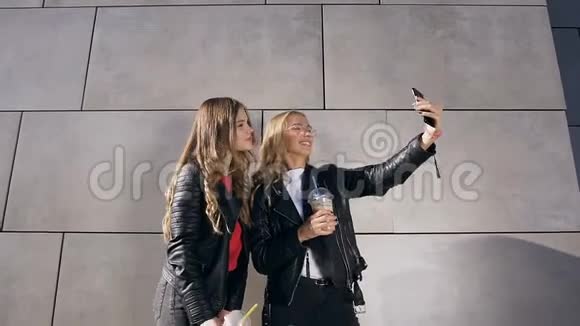 两个迷人的微笑高加索女孩在智能手机上自拍照片附近的灰色墙户外活动人朋友和视频的预览图
