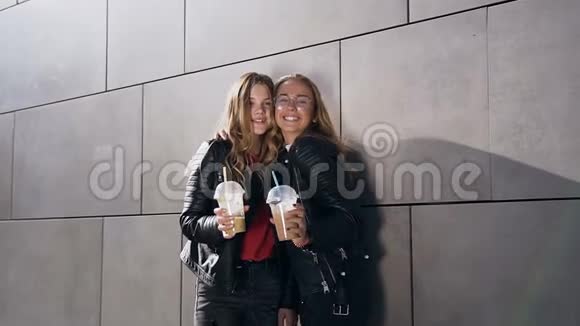 两个迷人的年轻女人拿着冰冷的鸡尾酒在夏天在现代建筑的灰色墙壁附近摆姿势室外最好视频的预览图
