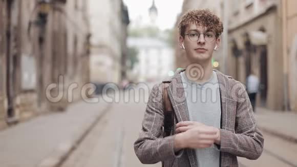 戴眼镜和戴耳机的年轻微笑的嬉皮士男子站在旧城街道背景下的画像视频的预览图
