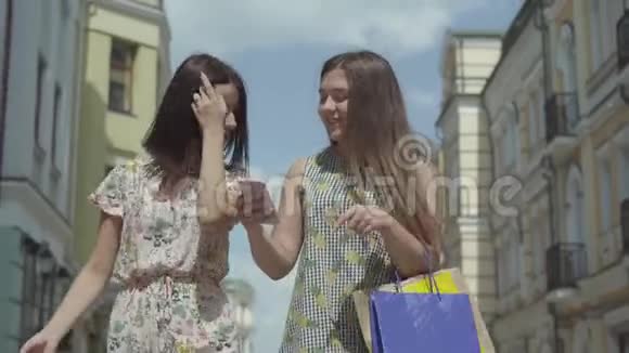 两位快乐的女朋友购物后拿着购物袋在漂亮的楼房前谈论新闻看着视频的预览图
