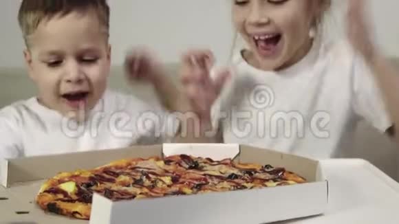 两个穿着白色t恤衫的可爱的孩子一个男孩和一个女孩打开一盒披萨享受美味视频的预览图