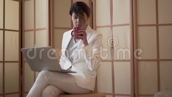 可爱的成年白种人女士喝着咖啡看着坐在家里的笔记本电脑屏幕女人揉着僵硬的脖子慢慢视频的预览图