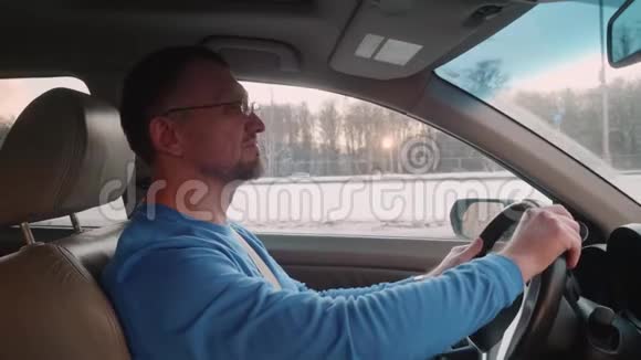 白色司机驾驶汽车在高速公路上疲惫而快乐的男人坐在车里对着音乐点头看着相机冬天日落视频的预览图