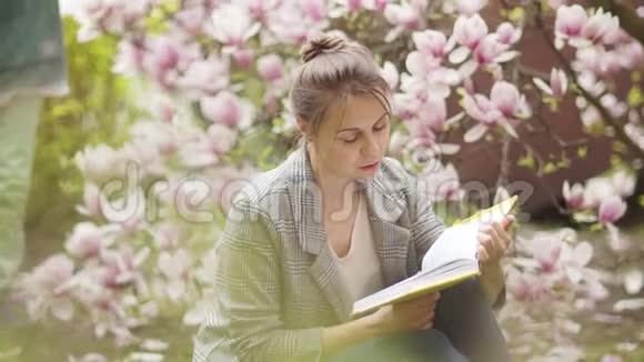 女人在一棵盛开的粉红色玉兰花的灌木背景上看书一个卷曲的儿子走到她面前给了一个蒲公英视频的预览图
