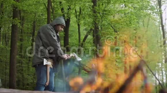 在野生落叶林的营地中在温暖的篝火附近与斧头和柴火一起工作的休闲强壮的人的缓慢运动视频的预览图