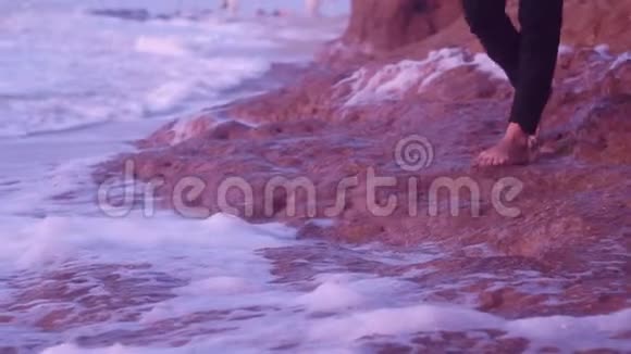一个年轻女孩的腿走在土石上海滩上日出时带泡沫的波浪视频的预览图