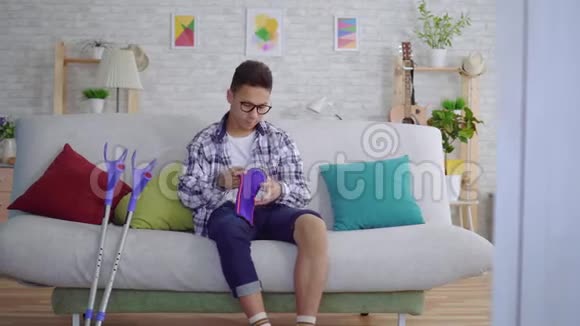 年轻的亚洲男人坐在沙发上用一条绷紧的弹力绷带绑在生病的膝盖上视频的预览图
