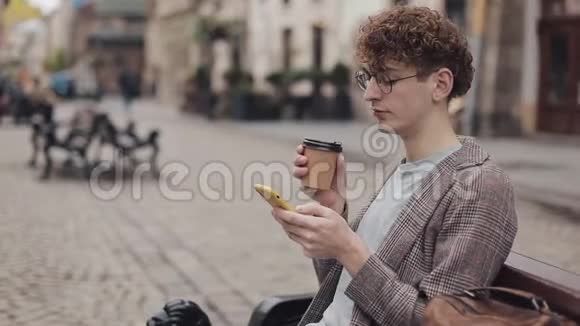 穿着夹克和眼镜的学生的侧视图喝咖啡一边浏览他的现代手机屏幕视频的预览图
